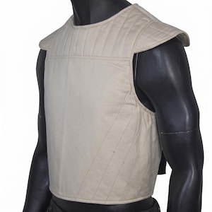Peut inclure: Un gilet beige matelassé avec épaulettes. Le gilet sans manches est fait d'une matière douce et rembourrée. Les coutures créent un motif de lignes verticales et diagonales. Le gilet est présenté sur un mannequin noir.