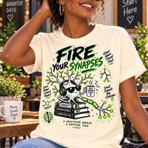 May include: A cream-colored short-sleeve t-shirt featuring a graphic of a cartoon neuron wearing headphones and sunglasses, sitting on a stack of books. The text reads: Fire Your Synapses, Curiosity, Neuroscience, A Brighter You, and A Healthier Brain A Happier You.