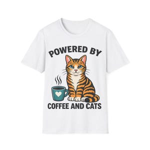 Op de afbeelding: Wit t-shirt met een afbeelding van een tijgerkat, een koffiekopje en de tekst "POWERED BY COFFEE AND CATS". De kat is oranje en zwart, met blauwe ogen. Het koffiekopje is turquoise met een hart.