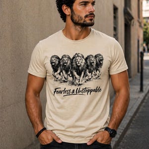 Op de afbeelding: Beige crewneck T-shirt met een afbeelding van vijf brullende leeuwen. De tekst "Fearless & Unstoppable" staat eronder in een scriptlettertype. Het T-shirt wordt door een persoon gedragen.