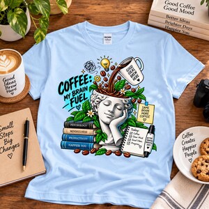 Op de afbeelding: Een lichtblauw t-shirt met een afbeelding van een klassiek standbeeldhoofd met een open bovenkant, waar koffie uit een mok in wordt gegoten. Het ontwerp bevat de tekst COFFEE: MY BRAIN FUEL, een stapel boeken, een notitieblok en koffiebonen.
