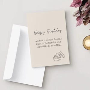 May include: A beige rectangular birthday greeting card with a white envelope. The card features black text that reads Happy Birthday, Another year older, but let's focus on the fact that your skin still looks incredible, and a line drawing of a jar.