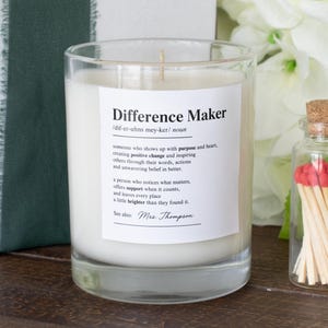 May include: A glass candle with the words "Difference Maker" and a definition on the label. Beside it is a small glass bottle filled with red-tipped matches. White flowers and a wrapped gift are in the background, creating a soft focus.