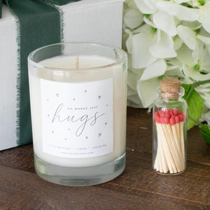 May include: A glass candle with the word "hugs" on the label, alongside a small glass bottle filled with red-tipped matches. A white gift box with a green ribbon and white flowers are also present.