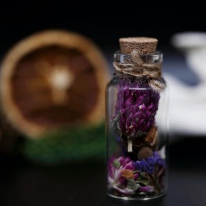 May include: A small, clear glass bottle filled with dried purple clover blossoms and small blue flowers. The bottle is sealed with a light brown cork stopper and features a piece of rustic twine tied around the neck of the glass container.