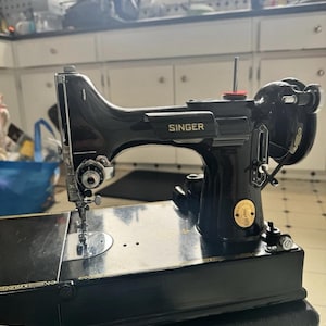 May include: A black Singer sewing machine with a glossy finish, featuring gold-coloured decals, a metal needle bar, a tension dial, and a circular emblem on the side. The machine has a horizontal spool pin with a red felt disc and a hand wheel.