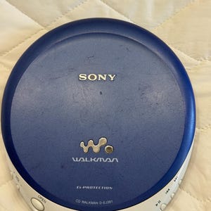 Può includere: Un lettore CD Sony Walkman blu e bianco. Il lettore CD portatile presenta il logo Walkman e il testo "G-PROTECTION" e "CD WALKMAN D-EJ361". Il dispositivo ha pulsanti per il volume, l'apertura e i controlli di riproduzione.