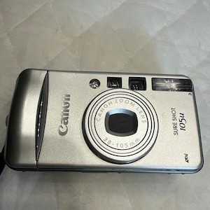May include: A silver Canon Sure Shot 105u point-and-shoot camera with a 38-105mm zoom lens. The front of the camera features the Canon logo, a flash unit, a viewfinder, and an autofocus sensor. The metallic finish has a smooth, rectangular design.