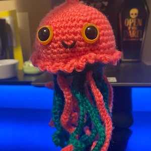 May include: A handmade crochet jellyfish ornament. The jellyfish is primarily coral-colored with large, black eyes outlined in yellow. Its tentacles are a mix of coral and green yarn, and it has a smiling face.