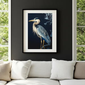 Op de afbeelding: Een ingelijste kunstdruk met een gedetailleerde illustratie van een blauwe reiger. De vogel is afgebeeld met blauwe, grijze en witte veren, een gele snavel en lange poten. Het kunstwerk staat tegen een donkere achtergrond.