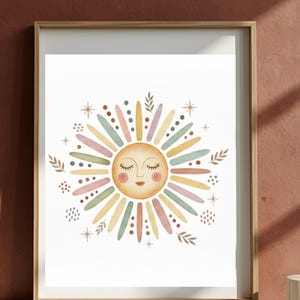 Op de afbeelding: Een ingelijste aquarelprint met een grillig zonontwerp. De zon heeft een lachend gezicht met gesloten ogen en is omgeven door kleurrijke stralen in pasteltinten van geel, roze, blauw en groen. Decoratieve sterren en bladeren completeren het kunstwerk.
