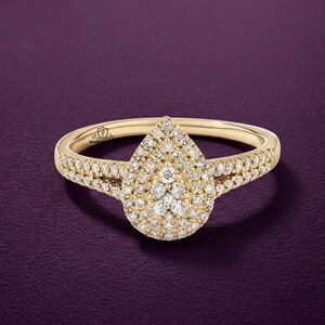 Peut inclure: Une bague en or avec un groupe de petits diamants en forme de poire au centre. La bande et les côtés de la forme de poire sont également incrustés de petits diamants. La bague est posée sur un fond violet foncé.