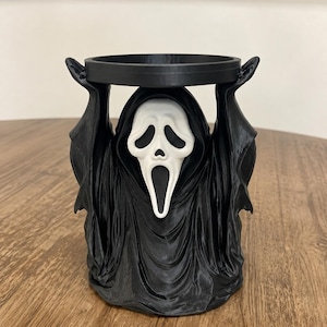 Peut inclure: Une figurine imprimée en 3D noire et blanche représentant un personnage encapuchonné avec un masque blanc au visage allongé et à la bouche ouverte. Les bras levés soutiennent une plateforme circulaire noire, formant un support fonctionnel.