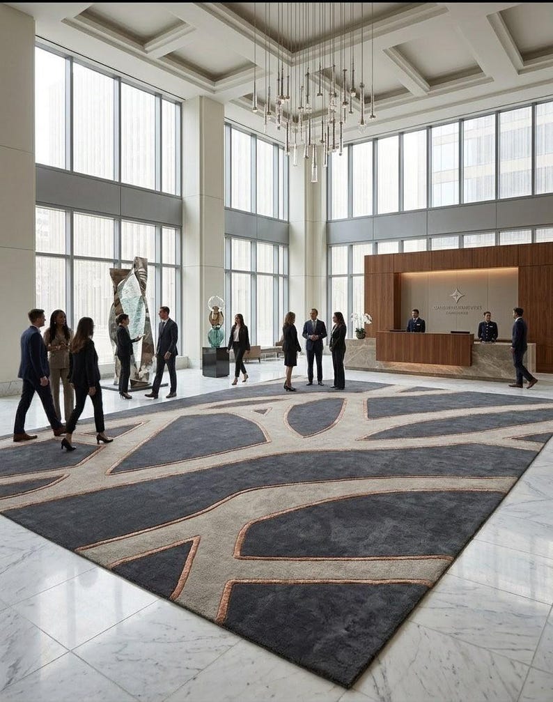 May include: A large, rectangular rug with a geometric design in a modern lobby. The rug features a dark gray background with light beige and copper-colored accents. The lobby has large windows, a high ceiling, and a reception desk.