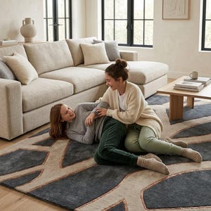 May include: A large, modern area rug with a geometric design in shades of gray, beige, and copper. The rug is placed in a living room setting with a beige sectional sofa and two people sitting on the rug.