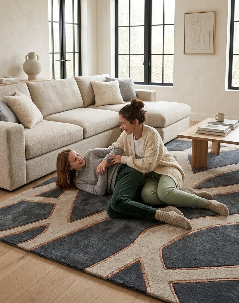 May include: A large, modern area rug with a geometric design in shades of gray, beige, and copper. The rug is placed in a living room setting with a beige sectional sofa and two people sitting on the rug.