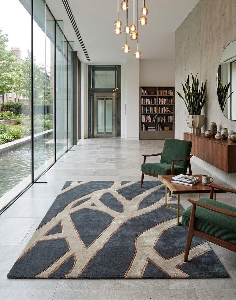 May include: A modern living room with a large, dark gray rug featuring a beige tree branch design. Two green armchairs with wooden frames and a small wooden table are in front of the rug. A large window and a decorative light fixture are also visible.