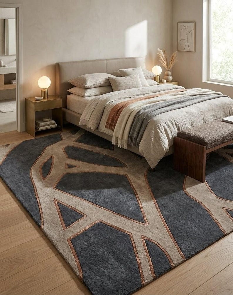 May include: A bedroom scene featuring a bed with neutral-toned bedding, a patterned rug, and a wooden bench. The rug has a geometric design in shades of gray, beige, and copper. Two small lamps sit on the nightstands.