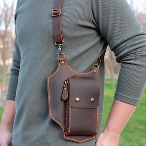 Könnte beinhalten: Eine braune Brusttasche aus Leder mit Schulterriemen. Die Tasche hat eine kleine Vordertasche mit Reißverschluss und zwei knopfartigen Verschlüssen in Messingfarbe. Die Tasche wird über der Schulter und über der Brust getragen.