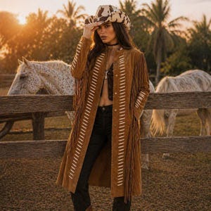 May include: A brown suede fringed coat with white stitching and a matching cowboy hat. The coat is long and open, with fringe along the sleeves and hem. The hat has a cowhide pattern. Two white horses stand behind a wooden fence.