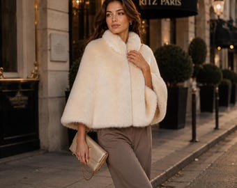 Op de afbeelding: Een crèmekleurige cape met een ronde kraag en korte mouwen. De vrouw draagt een bruine broek en draagt een gouden clutch met een kettingriem. De achtergrond bevat een gebouw met de tekst "RITZ PARIS".