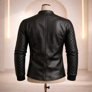 May include: Black leather jacket with quilted shoulder and sleeve detailing. The jacket has a stand-up collar and a tailored fit. The sleeves end with a black ribbed cuff. The jacket is displayed on a mannequin.