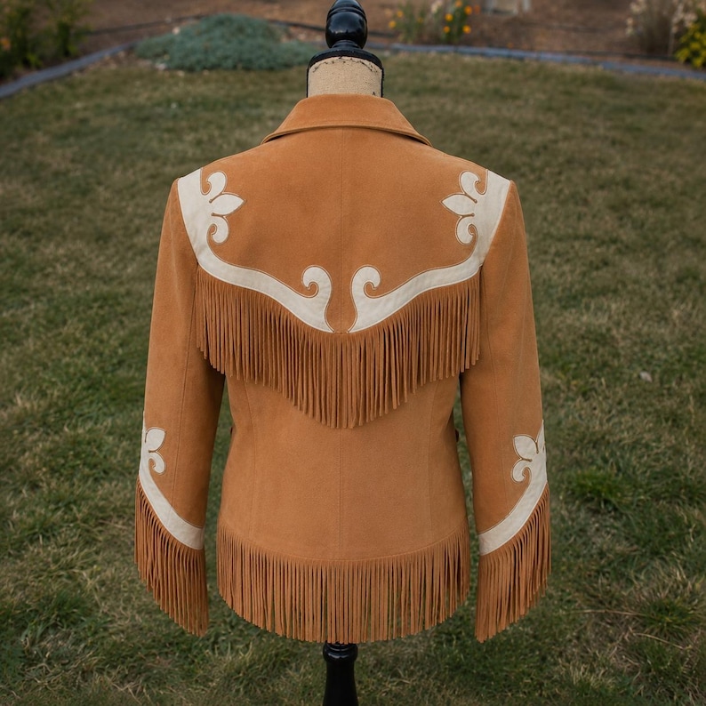 Op de afbeelding: Een beige su&egrave;de jas met lange franjes langs de zoom en mouwen. De jas is versierd met cr&egrave;mekleurige lederen overlays in een decoratief krulpatroon over het rugpand, die doorlopen langs de mouwen tot aan de boorden bij de polsen.