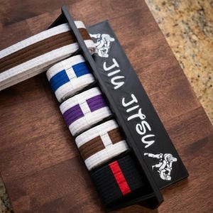 Może przedstawiać: Drewniany stojak ekspozycyjny z kilkoma pasami do Jiu Jitsu w różnych kolorach. Pasy obejmują brązowy, niebieski, fioletowy i czarny z czerwonym paskiem. Stojak jest czarny z białym napisem "Jiu Jitsu".
