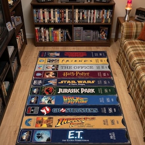 May include: A rectangular area rug featuring a graphic design of ten stacked VHS tape spines. The titles include Stranger Things, Friends, The Office, Harry Potter, Star Wars, Jurassic Park, Back to the Future, Ghostbusters, Indiana Jones, and E.T.