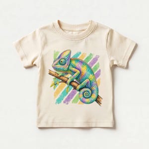May include: A cream-colored t-shirt featuring a graphic illustration of a multi-colored chameleon perched on a brown tree branch. The chameleon displays vibrant shades of blue, green, yellow, and purple, set against a background of diagonal stripes.