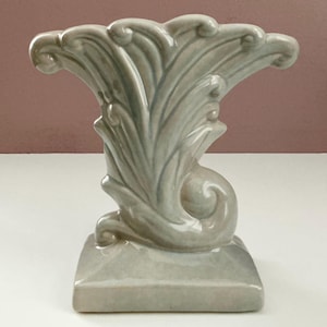 May include: A light grey ceramic vase with an ornate design. The vase has a square base and a flared top with a wave-like pattern. The height is approximately 18 cm.