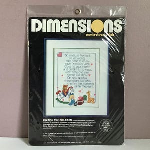 May include: A Dimensions counted cross stitch kit in a black package. The kit features a framed cross-stitch design with a poem and images of a teddy bear, rocking horse, and giraffe. The text reads "Cherish the Children."