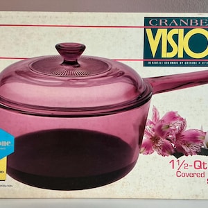 May include: A cranberry-colored Visions covered non-stick saucepan on a box. The glass saucepan has a handle and lid. The box has text that reads "Cranberry Visions" and "1 1/2-Qt./1.5L". Pink flowers are on the right.