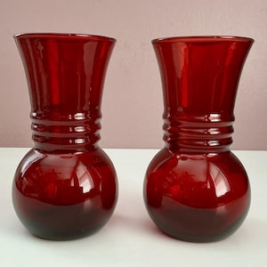 May include: Two ruby red glass vases. Each vase has a bulbous base, a cylindrical neck with horizontal ridges, and a flared top. The vases are made of a glossy, transparent glass.