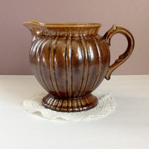 May include: A brown ceramic pitcher with a ribbed design, a curved handle, and a spout. The pitcher sits on a white lace doily. The pitcher has a glossy finish and is likely vintage.
