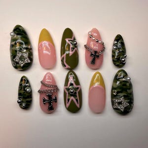 Könnte beinhalten: Eine Sammlung von zehn künstlichen Fingernägeln in verschiedenen Designs. Einige Nägel weisen ein Camouflage-Muster auf, andere haben rosa oder gelbe Basen mit dekorativen Elementen wie Sternen, Kreuzen und Ketten. Einige Nägel sind mit kleinen, silberfarbenen Perlen verziert.