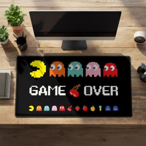 Puede incluir: Alfombrilla de escritorio rectangular negra con un diseño pixelado de personajes de Pac-Man y las palabras "GAME OVER" en blanco. El diseño incluye un Pac-Man amarillo, fantasmas naranjas, turquesas, rosas y rojos, y cerezas y fresas pixeladas.