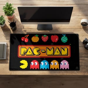 Puede incluir: Alfombrilla de escritorio rectangular negra con el logotipo de Pac-Man en amarillo y naranja, con frutas y fantasmas pixelados. La alfombrilla está colocada sobre un escritorio de madera, con un monitor de ordenador y plantas en el fondo.