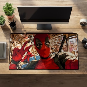 May include: Rectangular mousepad featuring a comic-style illustration of Deadpool making a peace sign. The red and black mousepad has a bus scene background with other characters. The word "FAIT" is visible.