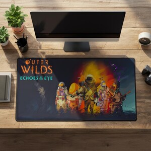 Pode incluir: Um tapete de mesa retangular preto com o texto "Outer Wilds Echoes of the Eye" e uma ilustração colorida de personagens em trajes espaciais. O tapete está em uma mesa de madeira com um monitor, plantas e um relógio.