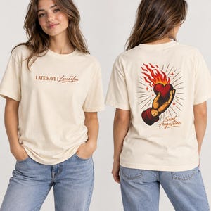 May include: A cream-coloured short-sleeve t-shirt featuring the text "LATE HAVE I Loved You" on the front. The back displays a graphic of a hand holding a flaming heart with the text "Saint Augustine" written in a script font below the illustration.