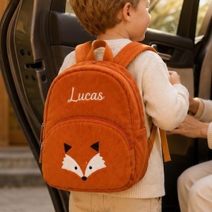 May include: A burnt orange corduroy backpack featuring a white embroidered fox face on the front pocket and the name Lucas in white script lettering. The backpack has adjustable shoulder straps and a top handle, with a textured fabric finish.
