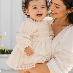 May include: A cream-coloured long-sleeved dress featuring a smocked bodice, a ruffled neckline, and intricate lace detailing along the hem. The garment is crafted from a textured fabric with gathered sleeves and a flared skirt silhouette.