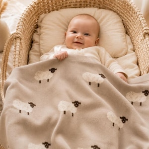 Könnte beinhalten: Eine hellbeige Strickdecke für Babys mit einem sich wiederholenden Muster aus kleinen weißen Schafen mit dunkelbraunen Köpfen und Beinen. Die Decke liegt über einem Baby, das in einem geflochtenen Weidenkorb mit einem cremefarbenen Kissen liegt.