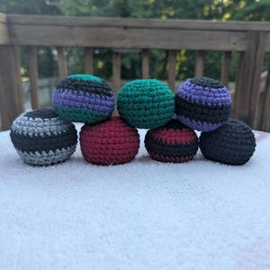May include: A collection of eight handmade crochet juggling balls in various colors. The balls are stacked on a white surface and feature stripes of black, gray, green, purple, and red. The balls are round and appear to be filled with a soft material.