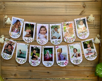 May include: A floral-themed photo banner with 12 pictures of a baby girl at different ages. Each picture is framed with a white border and has a number from 1 to 12 printed below it. The banner is strung together with twine and has small white tassels at each end.