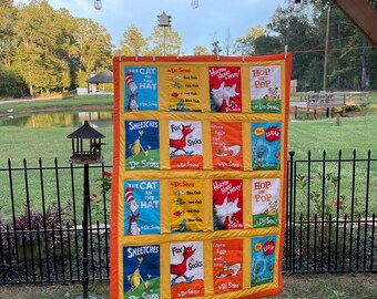 Puede incluir: Una colcha rectangular que presenta una cuadrícula de doce paneles de tela impresos con portadas de libros del Dr. Seuss, incluyendo The Cat in the Hat, One Fish Two Fish Red Fish Blue Fish, Horton Hears a Who, Hop on Pop, The Sneetches, Fox in Socks, Green Eggs and Ham y The Lorax, todo enmarcado por un borde de color naranja brillante.