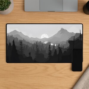Puede incluir: Una alfombrilla de escritorio rectangular con un diseño de paisaje en escala de grises. La obra de arte representa una cordillera, un bosque, un río y un sol poniente. Una persona está de pie en un acantilado. Un smartphone negro está a la derecha.