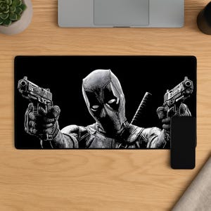 May include: A rectangular black desk mat featuring a high-contrast, monochromatic illustration of the character Deadpool. The character is depicted from the chest up, wearing a mask and holding two handguns pointed forward, with a sword hilt visible.