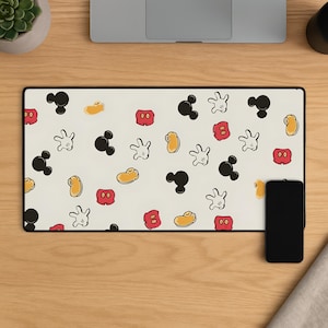 Puede incluir: Alfombrilla de ratón rectangular con fondo crema y un patrón de siluetas negras de Mickey Mouse, guantes blancos, pantalones cortos rojos y zapatos amarillos. Un smartphone negro descansa a la derecha.